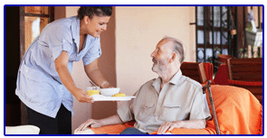 A woman serves a plate of food to an elderly man seated at a table, both smiling warmly at each other.